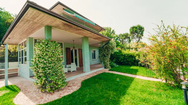 Modern house with a patio, green lawn, and surrounding trees in daylight.