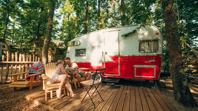 Forest camping with red and white trailer, two people sitting outside.
