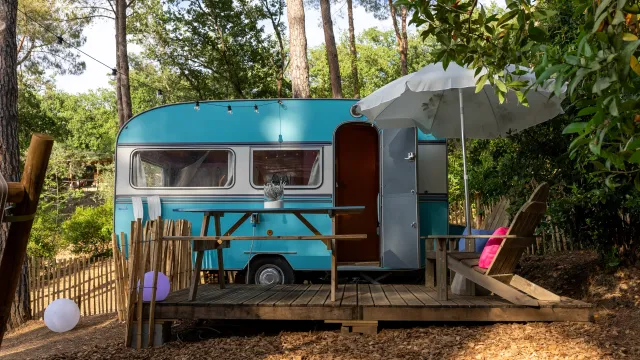 Blue vintage camper in a wooded setting with a deck, parasol, and chairs.