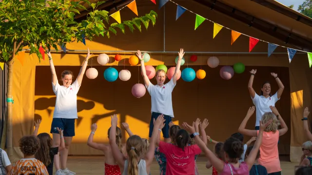 Children and instructors dance energetically under colorful bunting on a stage.