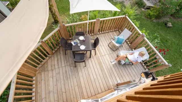 Sunlit wooden deck with a dining table, parasol, and a person lounging.