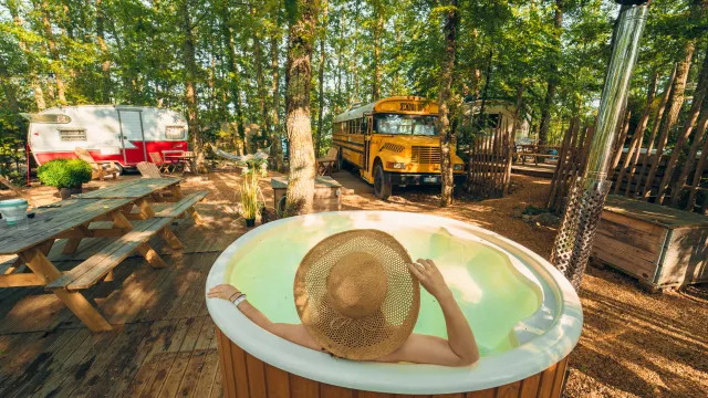Person in a hat relaxing in a forest hot tub, with a bus and camper nearby.