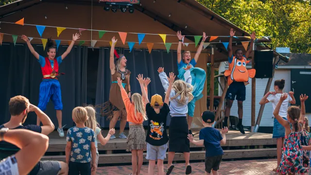 Children and performers on stage, colorful flags overhead, outdoors.