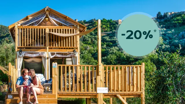 Wooden cabin with deck on a hillside; family sitting outside; clear blue sky.