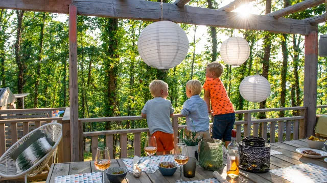 Children on a sunlit deck with lanterns, surrounded by trees.