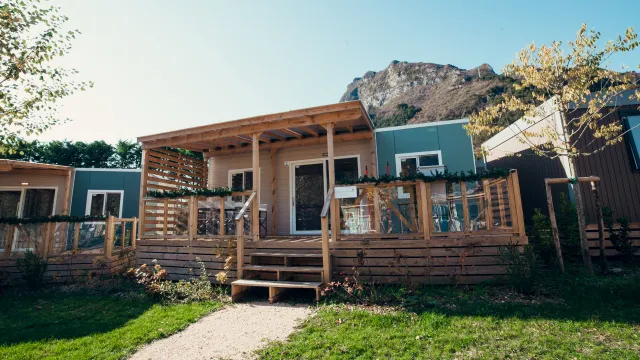 Cozy wooden cabin with porch, set against a mountain backdrop, surrounded by greenery.