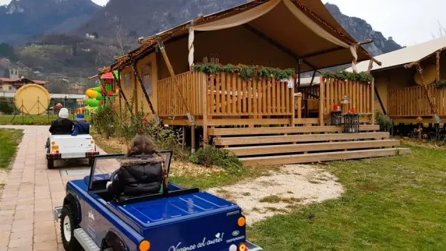 Children driving toy cars on a path near rustic cabins and mountains in the background.