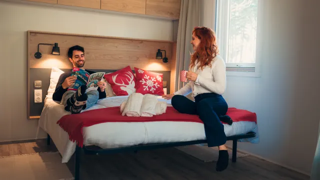 Man en vrouw praten op een bed in een warm verlichte kamer.