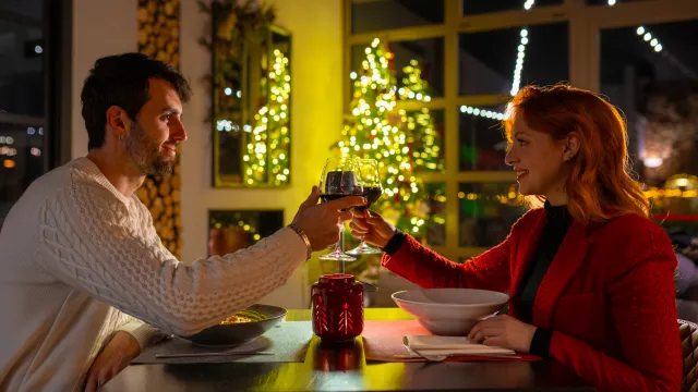 Couple toasting with wine at a cozy holiday dinner, lit Christmas tree in background.