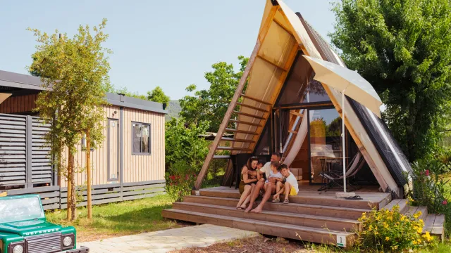 A-frame cabin with people sitting on the steps, surrounded by greenery and a small car nearby.
