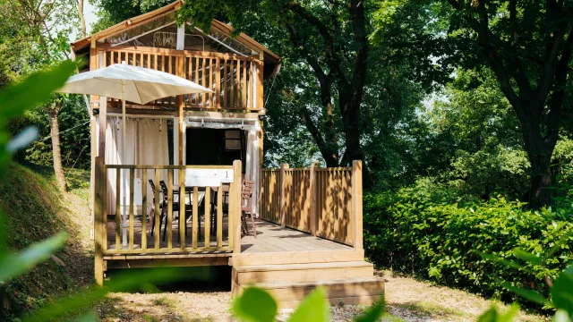 Wooden cabin with a balcony and umbrella, surrounded by lush greenery.