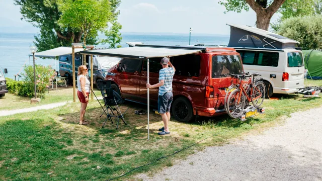 Camping scene with a red van near a lake, people setting up an awning, bikes on a rack.