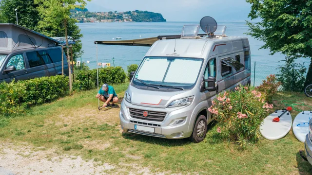Campervan parkeret ved en sø med en person, der sidder i nærheden, surfbræt og grønne omgivelser.