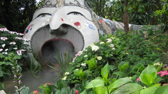 Large stone sculpture of face in garden, mouth open, surrounded by lush greenery.