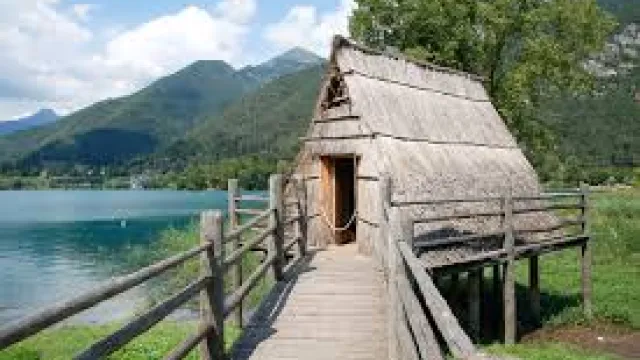 Wooden hut on a lake with mountain backdrop and clear sky.