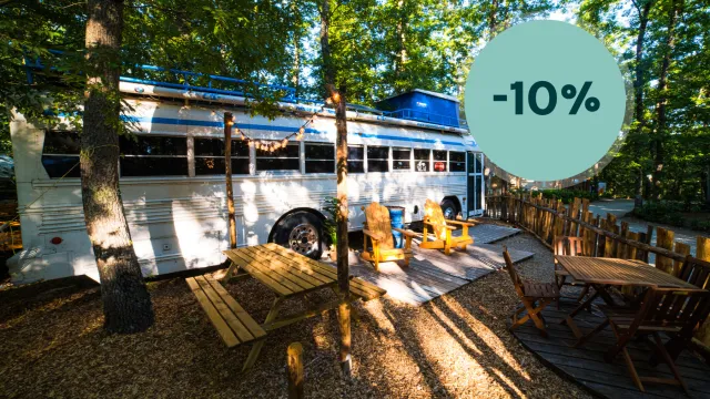 White bus converted into a campsite with chairs and tables, surrounded by trees.