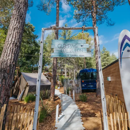 Camping site with tall trees, tents, blue bus, and surfboard under a clear sky.
