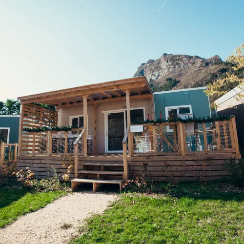 Cozy wooden cabin with porch, set against a mountain backdrop, surrounded by greenery.