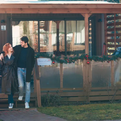 Couple smiling in front of a wooden cabin decorated with garlands.