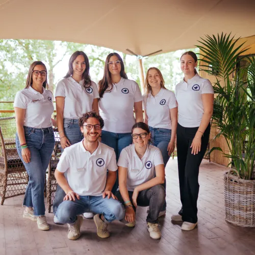 Group of seven people in white shirts, smiling on a wooden deck with potted plants.
