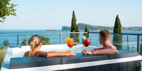 Couple relaxing in a hot tub with cocktails, overlooking a scenic lake and trees.