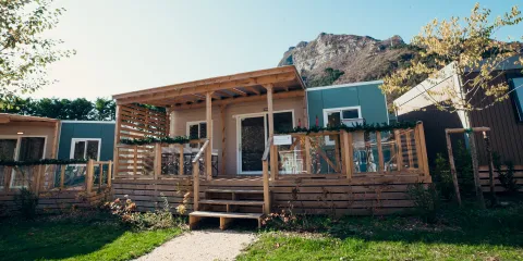 Cozy wooden cabin with porch, set against a mountain backdrop, surrounded by greenery.
