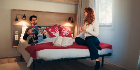 Man and woman chatting on a bed in a warmly lit room.