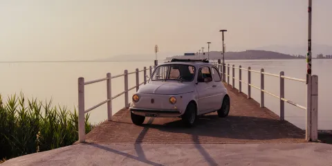 Vintage car on a pier at sunset, with calm water and soft pink sky.