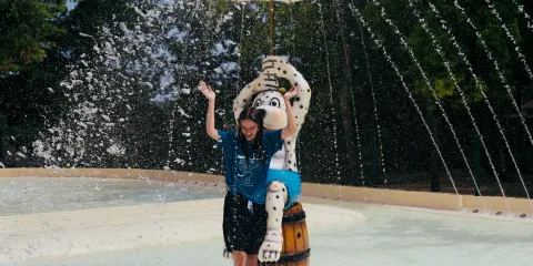 Person under a yellow umbrella fountain, surrounded by trees and blue sky.