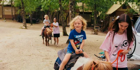 Child riding a pony led by a woman on a wooded path, other children on ponies behind.