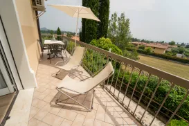 Sunny balcony with loungers, table, and umbrella, overlooking a garden and distant houses.