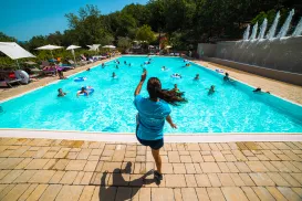 Person jumping into a crowded swimming pool on a sunny day, surrounded by trees.