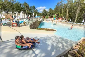 Two people on a waterslide in a sunny outdoor pool area with trees.