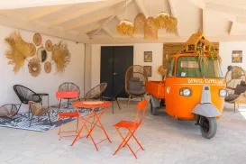 Orange vintage truck in a cozy patio with wicker decor and red metal chairs.