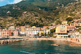 Coastal village with colorful buildings, boat in calm water, and mountain backdrop.