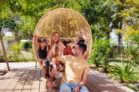 Group of friends smiling, sitting on a rattan swing in a sunny garden.