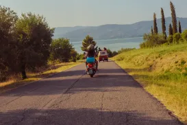 Person riding scooter on a country road toward a van, with trees and hills in the background.