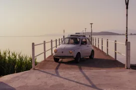 Small vintage car on a pier at sunset, with calm water and mountains in the background.