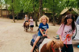 Child riding a pony led by a woman on a wooded path, other children on ponies behind.