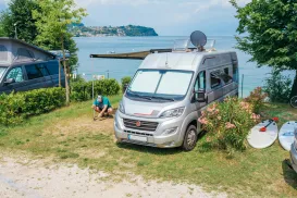 Camper van parked by a lake, man relaxing nearby, sunny day.