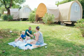Family picnicking on a grassy field near canvas-covered huts.