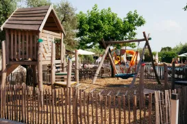 Wooden playground with swings and slide, surrounded by a fence and trees.
