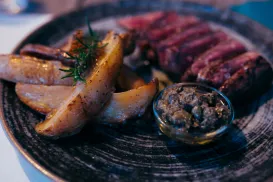 Sliced steak with potato wedges and sauce on a dark plate.
