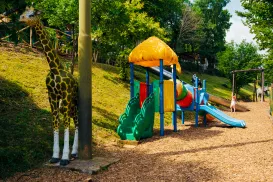 Playground with colorful slide, giraffe statue, and green trees.