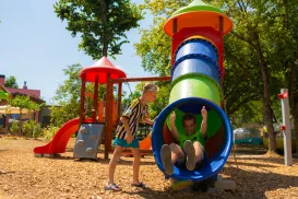 Colorful playground slide, child exiting, another child waiting on a sunny day.