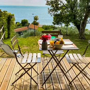 Deck with table, chairs, flowers, and breakfast, overlooking a lake and trees.