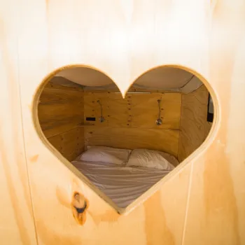 Cozy wooden nook with a heart-shaped window, showing a bed with pillows.