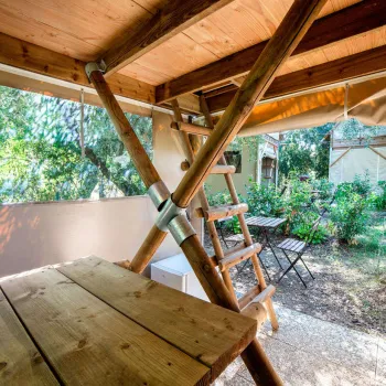 Wooden loft interior with ladder, opening to garden with chairs and trees.