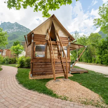 Wooden cabin with tent roof surrounded by trees, brick pathway and mountain backdrop.