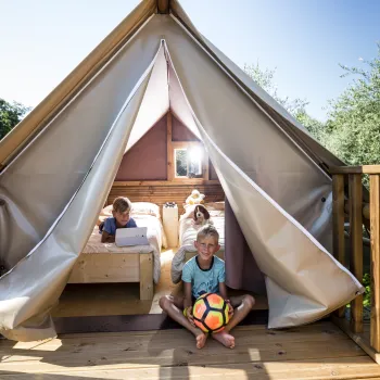 Kids sit in a wooden tent structure, one holding a colorful soccer ball, under a clear blue sky.